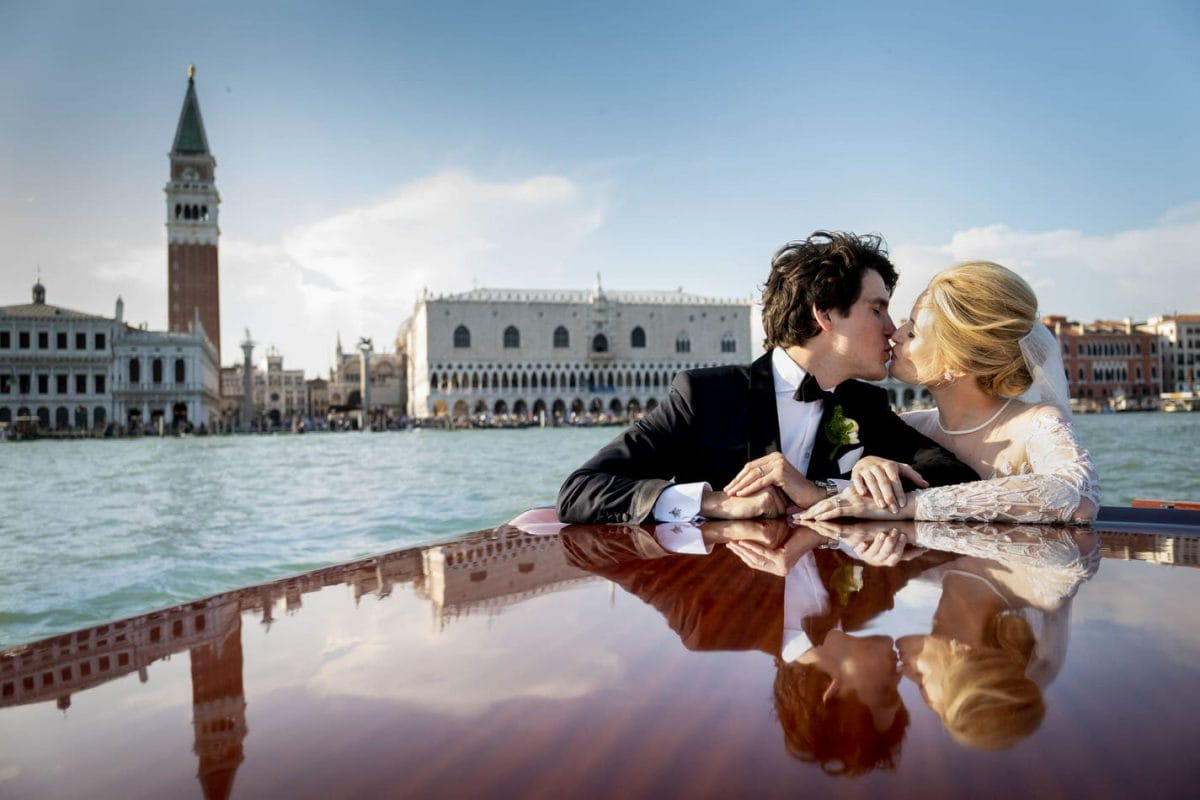 Wedding couple on a water taxi in Venice, Italy, kissing with historic landmarks in the background.
